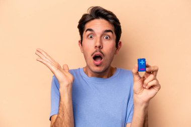 Beyazlı genç bir adam bej arka planda izole edilmiş bir bateri tutuyor. Şaşırmış ve şok olmuş..