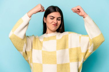 Mavi arka planda izole edilmiş genç beyaz kadın güçlü bir hareket sergiliyor kollarıyla, kadınsı gücün sembolü.