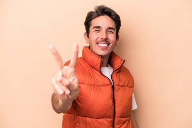 Beyaz tenli genç adam bej arka planda izole edilmiş neşeli ve kaygısız parmaklarıyla barış sembolü gösteriyor..
