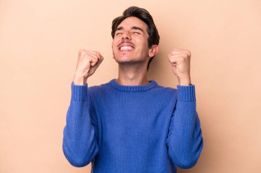 Beyazlı genç adam bej arka planda izole edilmiş zafer, tutku ve coşkuyu kutluyor, mutlu bir ifade..