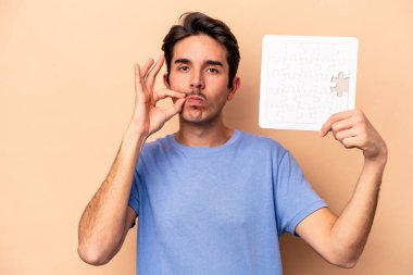 Beyazlı genç bir adam bej arka planda izole edilmiş bir yapboz tutuyor ve parmakları dudaklarında bir sır saklıyor..