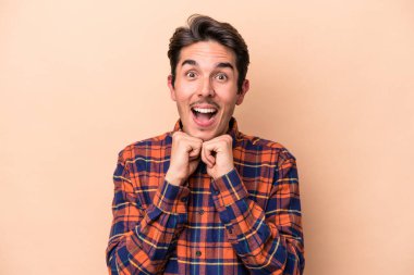 Beyaz tenli genç adam bej arka planda izole edilmiş şans için dua ediyor, şaşırmış ve ağzını açıp önüne bakıyor..