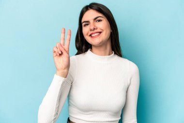 Mavi arka planda izole edilmiş genç beyaz kadın iki numarayı parmaklarıyla gösteriyor..