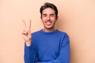 Beyazlı genç bir adam bej arka planda izole edilmiş. İki numarayı parmaklarıyla gösteriyor..