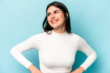 Mavi arka planda izole edilmiş genç beyaz kadın mutlu bir şekilde gülüyor ve ellerini karnının üstünde tutarak eğleniyor..