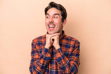 Beyaz tenli genç adam bej arka planda izole edilmiş ellerini çenesinin altında tutuyor, mutlu bir şekilde yana bakıyor..