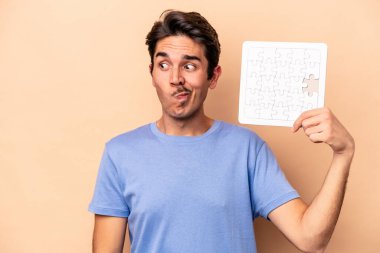 Kafkasyalı genç bir adam bej arka planda izole edilmiş bir yapboz tutuyor. Kafası karışmış ve kendinden emin değil..