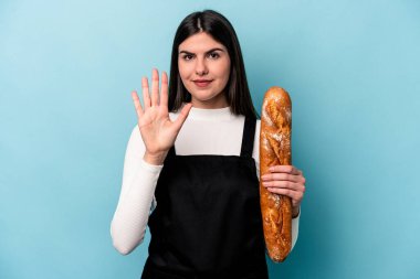 Genç fırıncı Kafkasyalı, mavi arka planda izole edilmiş bir somun ekmek tutuyor. Beş numarayı parmaklarıyla gülümsüyor..