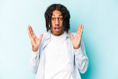 Genç Afrikalı Amerikalı bir adam mavi arka planda izole edilmiş. Şaşırmış ve şok olmuş..
