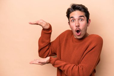 Beyazlı genç bir adam bej arka planda izole edilmiş. Elleri arasında bir kopya alanı tutarken şok olmuş ve hayrete düşmüş..