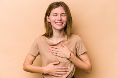 Bej arka planda izole edilmiş genç İngiliz kadın mutlu bir şekilde gülüyor ve ellerini karnının üstünde tutarak eğleniyor..