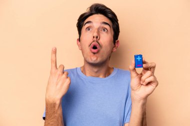 Beyazlı genç bir adam bej arka planda izole edilmiş bir bateriyi tutuyor ve ağzı açık bir şekilde yukarı bakıyor..