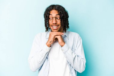 Mavi arka planda izole edilmiş genç Afrikalı Amerikalı ellerini çenesinin altında tutuyor ve mutlu bir şekilde yan tarafa bakıyor..