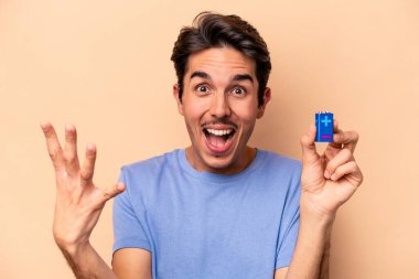 Beyazlı genç beyaz adam bej arka planda izole edilmiş bir bateriyi tutuyor. Hoş bir sürpriz alıyor, heyecanlı ve el kaldırıyor..