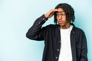 Genç Afrikalı Amerikalı adam mavi arka planda izole edilmiş, uzaklara bakıyor ve elini alnına koyuyor..