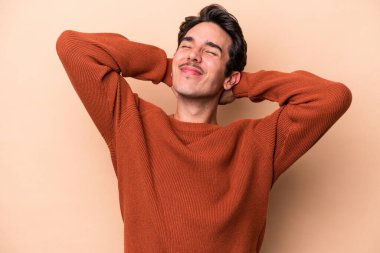 Beyazlı genç bir adam bej arka planda izole edilmiş. Kendine güveniyor, elleri başının arkasında..