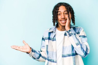 Mavi arka planda izole edilmiş genç Afro-Amerikalı bir adam avuç içinde fotokopi alanı tutuyor, el üstünde tutuyor. Şaşırmış ve mutlu..