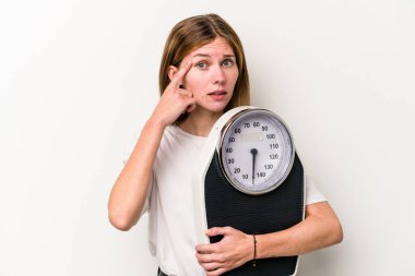 Beyaz arka planda izole edilmiş bir tartı tutan genç İngiliz kadın işaret parmağıyla yaptığı hayal kırıklığını gösteriyor..