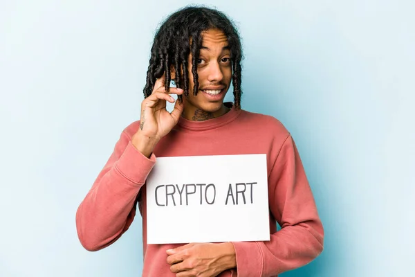 Young African american man holding crypto art placard isolated on blue background trying to listening a gossip.