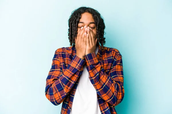 Genç Afrikalı Amerikalı bir adam mavi arka planda izole edilmiş bir şekilde bir şeylere gülüyor, elleriyle ağzını kapatıyor..