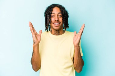 Mavi arka planda izole edilmiş genç Afro-Amerikalı adam yüksek sesle gülüyor ve elini göğsüne koyuyor..