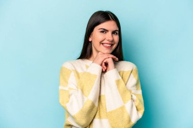 Mavi arka planda izole edilmiş genç beyaz kadın ellerini çenesinin altında tutuyor, mutlu bir şekilde yana bakıyor..