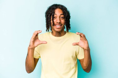 Genç Afrikalı Amerikalı adam mavi arka planda izole edilmiş. Elinde avuç içi olan bir şey var. Kameraya teklif ediyor..