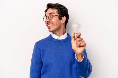 Beyaz arka planda izole edilmiş bir ampul tutan genç beyaz bir adam gülümseyen, neşeli ve keyifli bir kenara bakar..