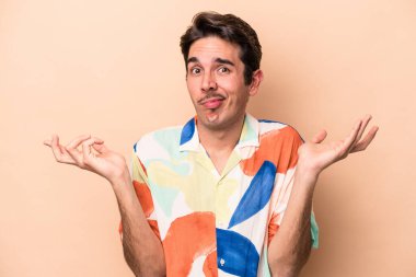 Beyazlı genç bir adam bej arka planda izole edilmiş sorgulama sırasında omuz silkiyor ve şüphe duyuyor..