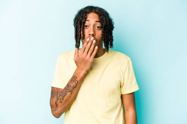 Genç Afrikalı Amerikalı bir adam mavi arka planda izole edilmiş. Şok olmuş, ağzı ellerle kaplanmış, yeni bir şey keşfetmeye hevesli..