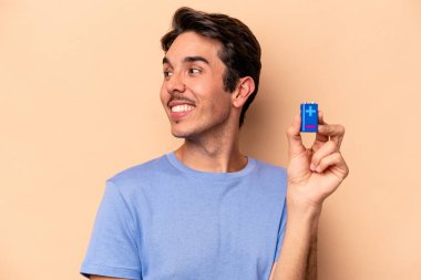 Beyaz tenli genç adam bej arka planda izole edilmiş bir bateriyi tutuyor. Gülümsüyor, neşeli ve keyifli görünüyor..
