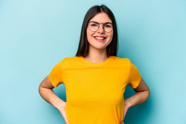 Kafkasyalı genç bir kadın mavi arka planda izole edilmiş. Kendine güvenen, ellerini kalçasında tutan..