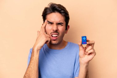 Beyazlı genç bir adam bej arka planda izole edilmiş bir bateriyi tutuyor ve işaret parmağıyla yaptığı hayal kırıklığı jestini gösteriyor..