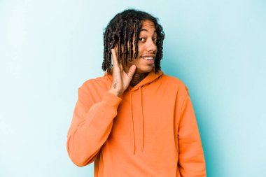 Genç Afrikalı Amerikalı bir adam mavi arka planda izole edilmiş bir şekilde bir dedikoduyu dinlemeye çalışıyor..