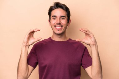 Beyaz tenli genç adam bej arka planda izole edilmiş. Elinde avuç içi olan bir şeyle kameraya sunuyor..