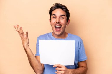 Beyazlı genç bir adam bej arka planda izole edilmiş bir pankart tutuyor. Hoş bir sürpriz alıyor, heyecanlı ve el kaldırıyor..