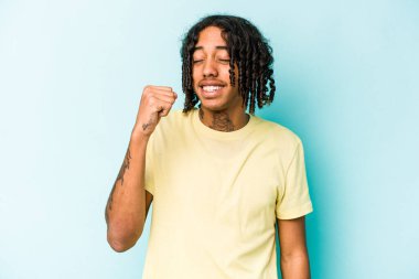 Genç Afrikalı Amerikalı bir adam mavi arka planda izole edilmiş zafer, tutku ve coşkuyu kutluyor, mutlu bir ifade..