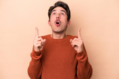 Beyazlı genç bir adam bej arka planda izole edilmiş. Ağzı açık, üst tarafı işaret ediyor..