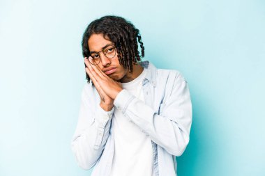 Genç Afrikalı Amerikalı bir adam mavi arka planda izole edilmiş esniyor ve ağzı eliyle kapalı yorgun bir jest yapıyor..