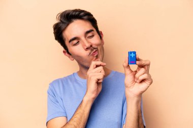 Beyazlı genç bir adam bej arka planda izole edilmiş bir bateriyi tutuyor şüpheli ve şüpheci bir ifadeyle yana bakıyor..