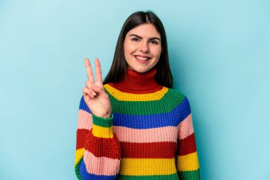 Mavi arka planda izole edilmiş genç beyaz kadın zafer işareti gösteriyor ve gülümsüyor..
