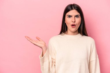 Pembe arka planda izole edilmiş genç beyaz kadın, avuç içi fotokopi alanını elinde tutmaktan etkilenmiş..