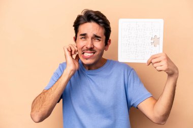 Kafkasyalı genç bir adam bej arka planda izole edilmiş bir yapboz tutuyor kulakları elleriyle kapatıyor..