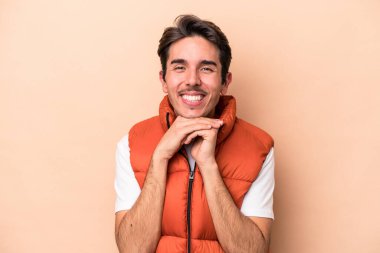 Beyaz tenli genç adam bej arka planda izole edilmiş ellerini çenesinin altında tutuyor, mutlu bir şekilde yana bakıyor..