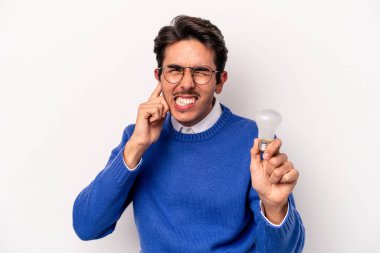 Beyaz arka planda izole edilmiş ampul tutan genç beyaz bir adam kulaklarını elleriyle kapatıyor..