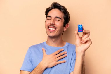 Beyazlı genç bir adam bej arka planda izole edilmiş bir bateriyi tutuyor kahkahalar atıyor ve elini göğsüne koyuyor.