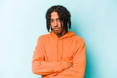 Mavi arka planda izole edilmiş, hoşnutsuzca kaşlarını çatmış, kollarını kavuşturmuş genç Afrikalı Amerikalı bir adam..
