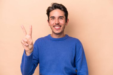 Beyaz tenli genç adam bej arka planda izole edilmiş neşeli ve kaygısız parmaklarıyla barış sembolü gösteriyor..