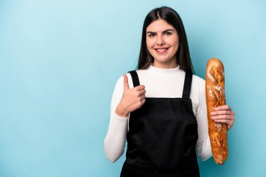 Genç fırıncı Kafkas elinde bir somun ekmekle mavi arka planda izole edilmiş gülümsüyor ve başparmağını kaldırıyor.