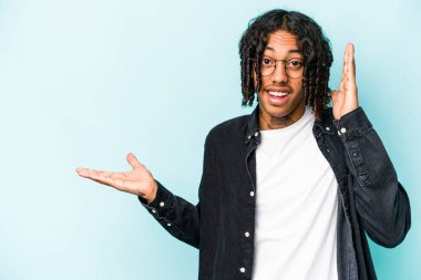 Mavi arka planda izole edilmiş genç Afro-Amerikalı bir adam avuç içinde fotokopi alanı tutuyor, el üstünde tutuyor. Şaşırmış ve mutlu..